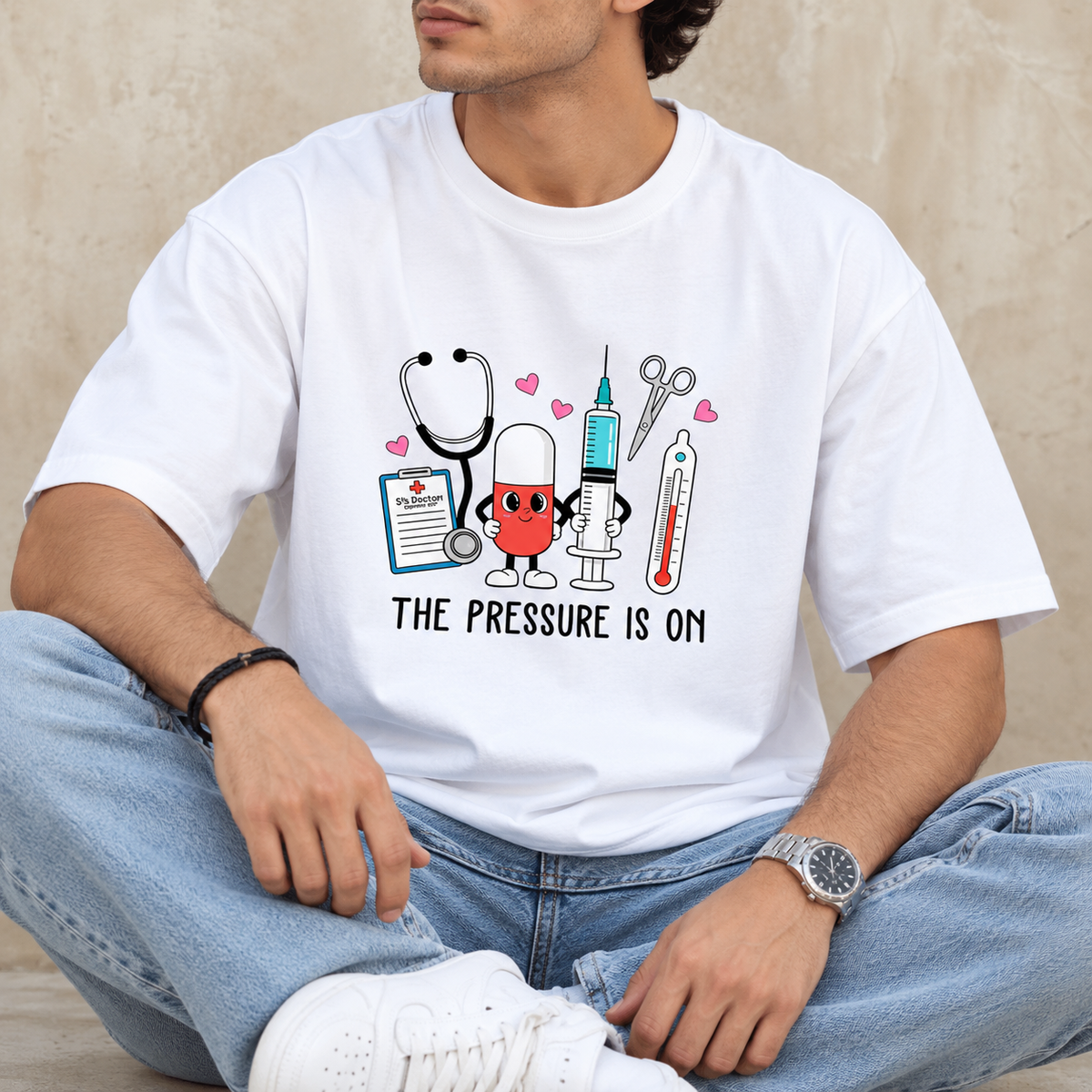 Man wearing a white graphic T-shirt featuring colourful medical illustrations, pink hearts and the text “THE PRESSURE IS ON.”
