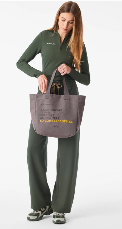 Woman wearing dark green medical scrubs and carrying a taupe-grey tote bag with medical-themed lettering in grey and yellow.