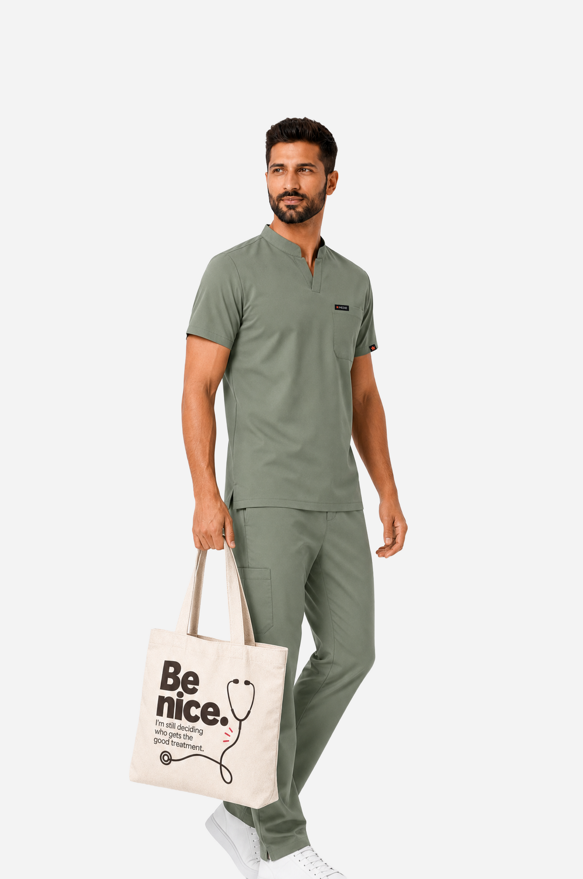 Male healthcare professional in sage-green scrubs carrying a cream tote bag printed with “Be nice. I’m still deciding who gets the good treatment” and a black stethoscope illustration.
