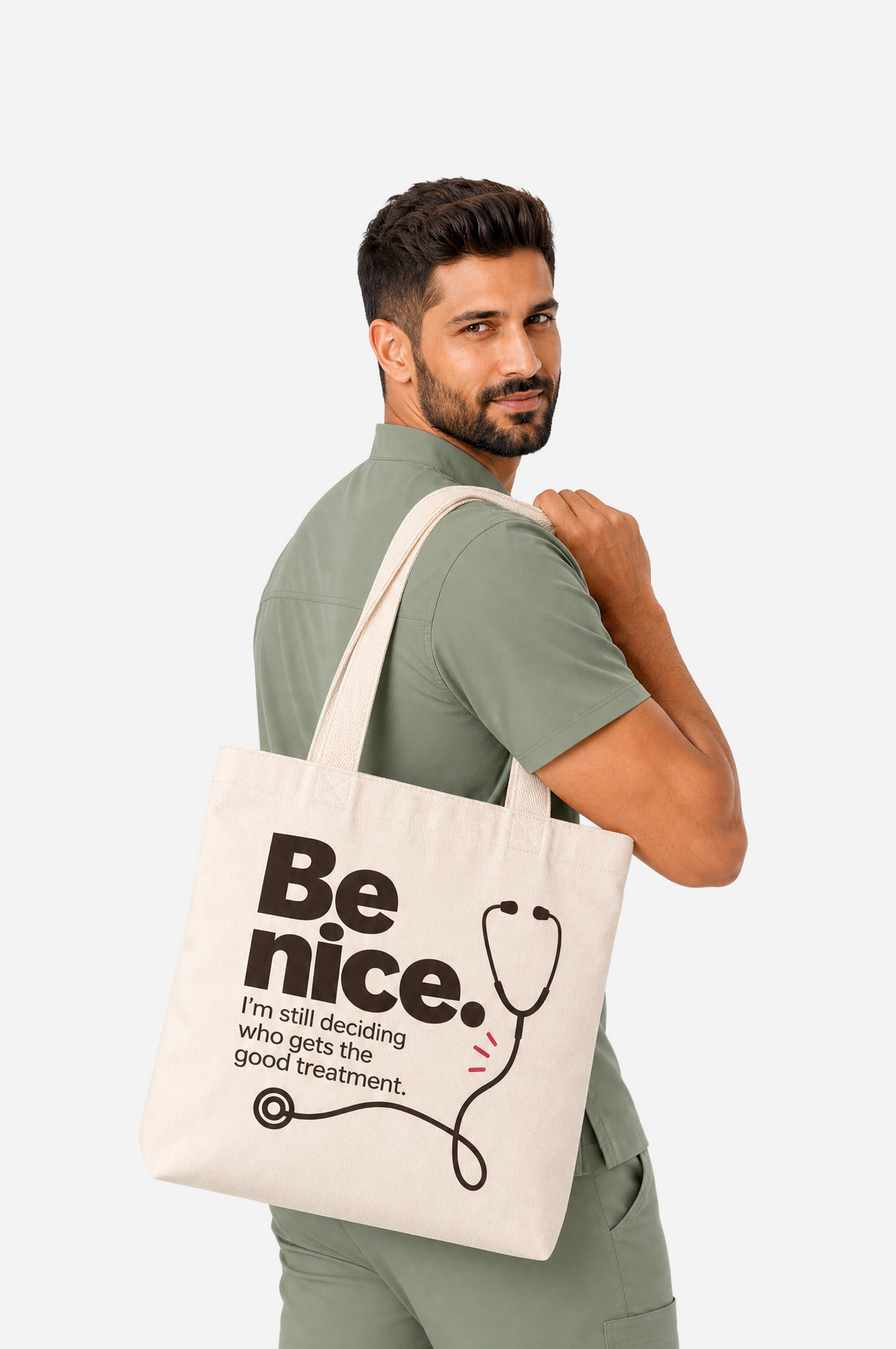 Male healthcare professional in sage-green scrubs carrying a cream tote bag printed with “Be nice. I’m still deciding who gets the good treatment” and a black stethoscope illustration.