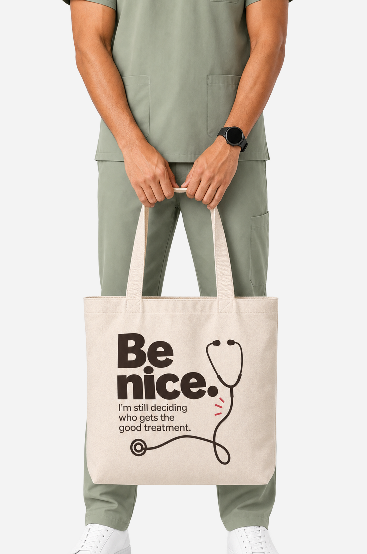 Male healthcare professional in sage-green scrubs carrying a cream tote bag printed with “Be nice. I’m still deciding who gets the good treatment” and a black stethoscope illustration.